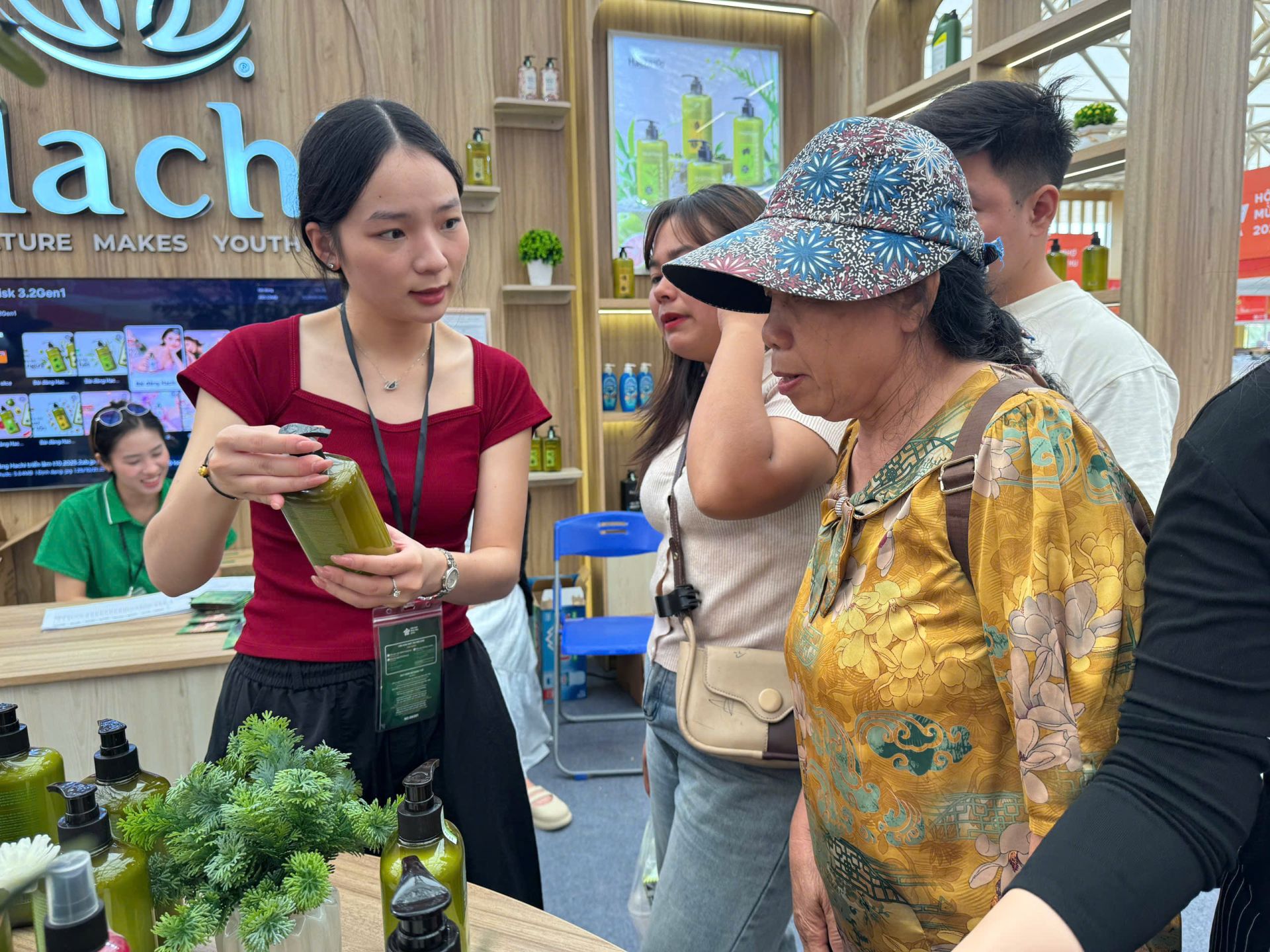 Nhân viên giới thiệu sản phẩm dầu gội chiết xuất thiên nhiên cho khách hàng lớn tuổi tại gian hàng Hachi.